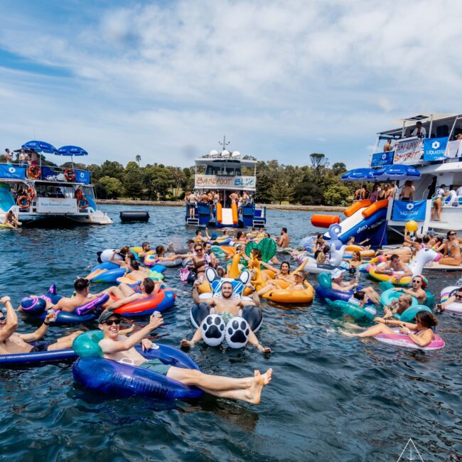 A group of people enjoy a sunny day on a lake, floating on colorful inflatable tubes and pool floats. Boats with slides are anchored nearby, and trees line the shore in the background. The scene is festive and lively.