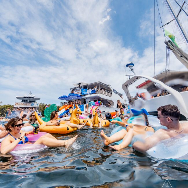 A group of people enjoying a sunny day on the water, lounging on colorful inflatable floats. Boats are anchored in the background. The sky is partly cloudy, creating a lively and relaxed atmosphere.