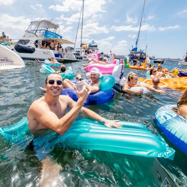 A group of people floating on inflatable rafts in a sunny body of water, surrounded by several boats. Some are smiling and posing for the camera, with bright blue skies overhead and a vibrant, lively atmosphere.