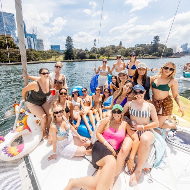 A group of women in swimsuits are gathered on a boat deck, posing and smiling. They're surrounded by water, with city buildings and trees in the background. Some hold drinks and sit on inflatable floats. It's a sunny day.