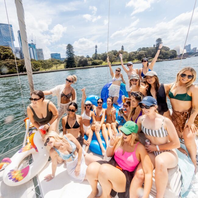 A group of people enjoying a sunny day on a sailboat. They are wearing swimsuits, sunglasses, and summer hats, some holding drinks. The backdrop includes city skyscrapers, a body of water, and trees under a partly cloudy sky.