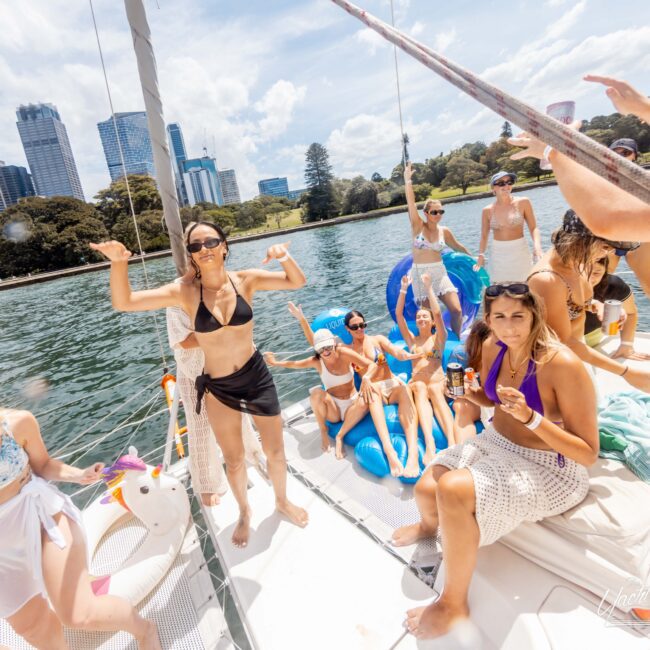 A group of people in swimsuits are enjoying a sunny day on a boat. Some are standing, dancing, and holding drinks. Inflatable pool toys and skyscrapers are visible in the background. The water glistens under a partly cloudy sky.