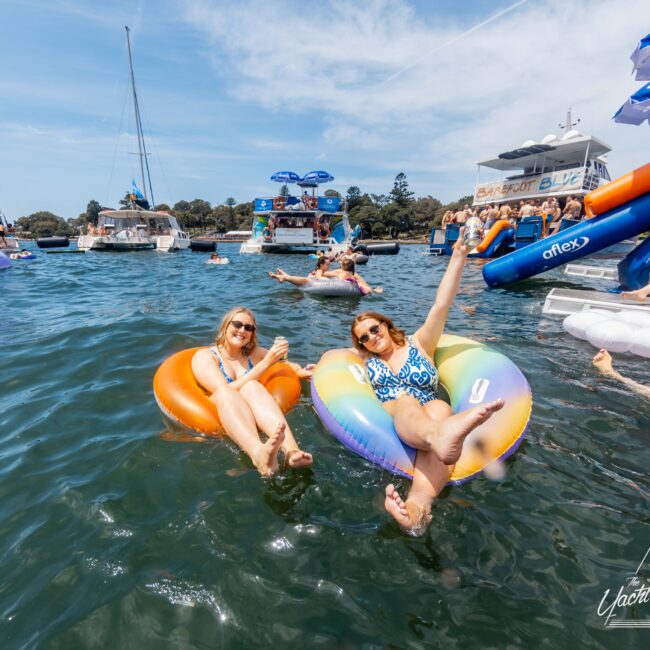 Two people relax on inflatable tubes in a lake, surrounded by boats and other floaters. One person waves cheerfully. The scene is lively, with clear blue skies and people enjoying the water.