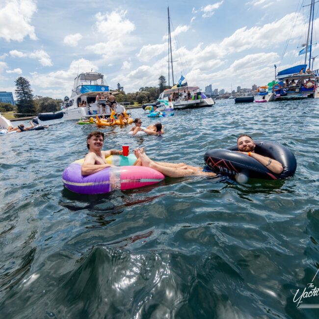 People are lounging on inflatable tubes in a vibrant, sunny harbor filled with boats. They are holding drinks and smiling, surrounded by others enjoying the water. A mix of sailboats and motorboats can be seen in the background under a partly cloudy sky.