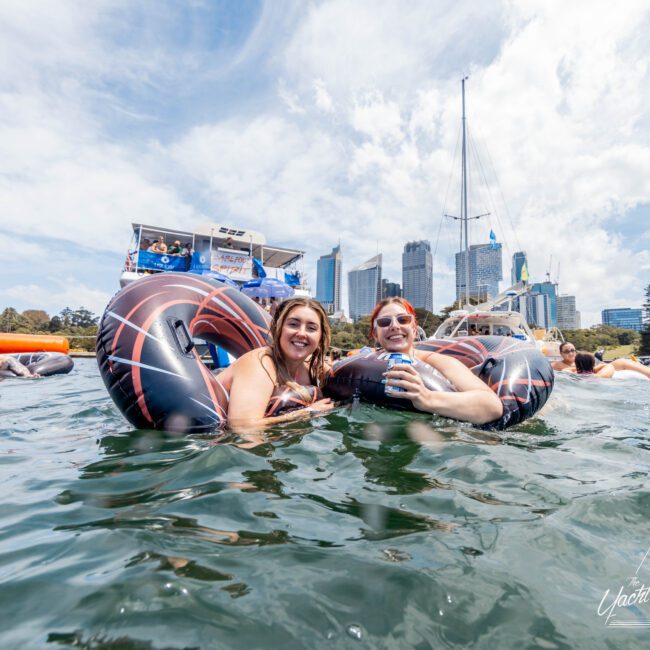 Two people smiling and floating in a pool on large inflatable rings. They are surrounded by a few others who are also swimming or floating. Boats and a cityscape with tall buildings are visible in the background under a partly cloudy sky.