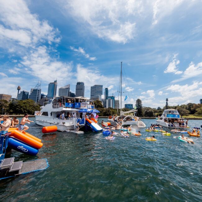 Boats and people in colorful floats enjoy a sunny day on the water with a city skyline in the background. The scene is lively with several boats anchored close to each other and people swimming and relaxing.