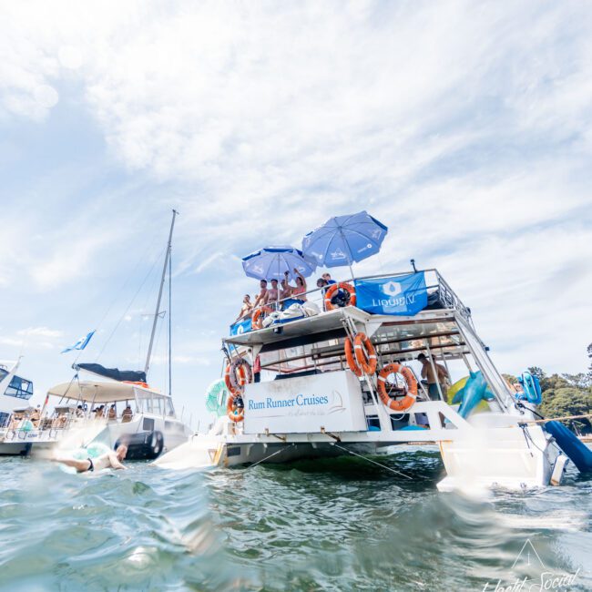 A cruise boat called "Rum Runner Cruises" is anchored on a sunny day. People relax on the upper deck under blue umbrellas. Nearby boats are visible, and one person is in the water. The sky is bright with scattered clouds.