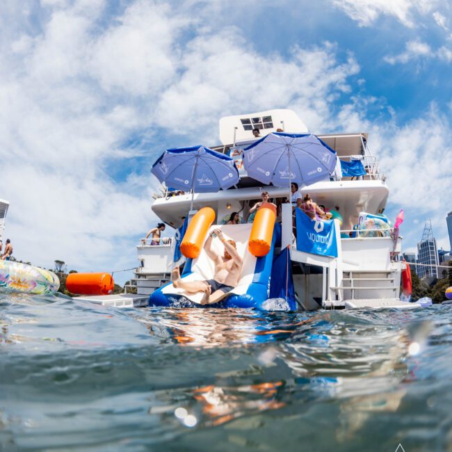 A person relaxes on a floating lounge chair with orange armrests in the water. Behind them is a two-story boat with blue umbrellas and people on board. The sky is mostly clear with some clouds.