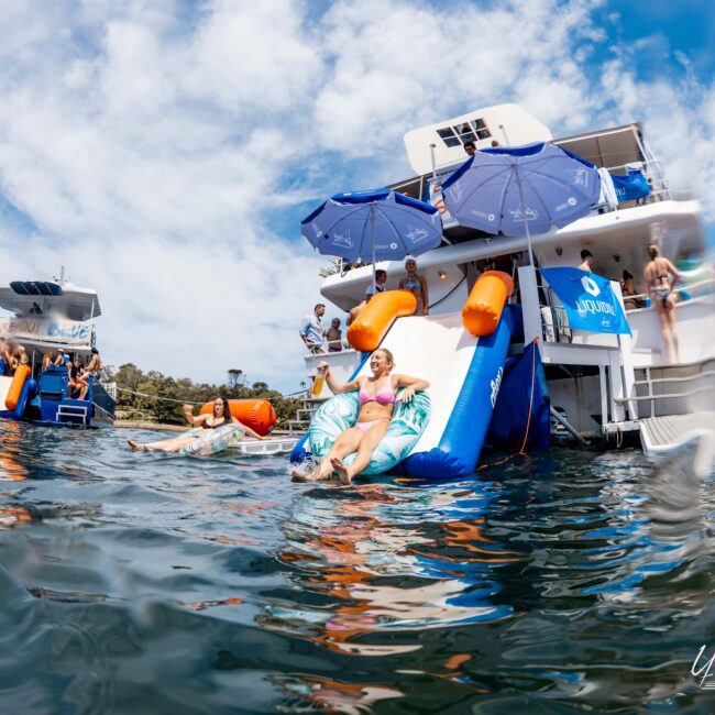 A group of people enjoying a sunny day on a yacht, with some relaxing on inflatable floats and others using a water slide attached to the boat. Bright blue sky and city buildings visible in the background.