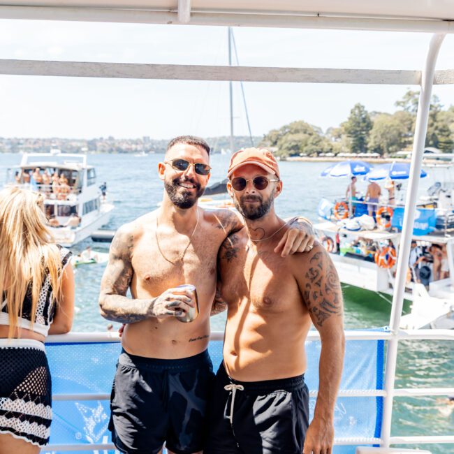 Two shirtless men in sunglasses stand on a boat, smiling and holding drinks. They are surrounded by other boats and people in swimwear. The scene is sunny, with trees and water in the background.