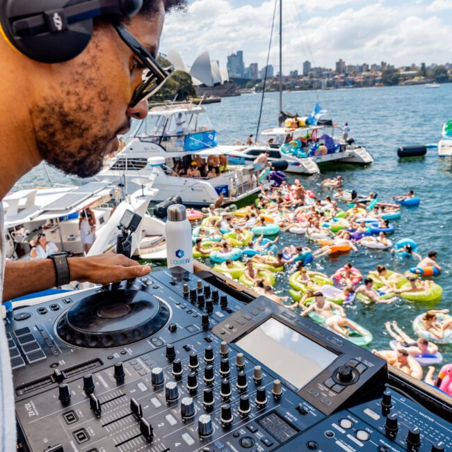 DJ with headphones performs at an outdoor party on a yacht. Crowd in pool floats enjoy the music on the water. City skyline and boats visible in the background under a clear, sunny sky.