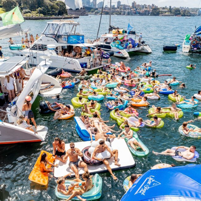 A lively scene on the water with many people on colorful inflatables gathered between boats. The sky is partly cloudy, and a city skyline is visible in the background. The atmosphere is festive and relaxed.