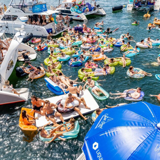 Crowded party scene on water with people lounging on colorful inflatables. Boats are anchored nearby, with a large blue umbrella in the foreground. The atmosphere is lively and festive.