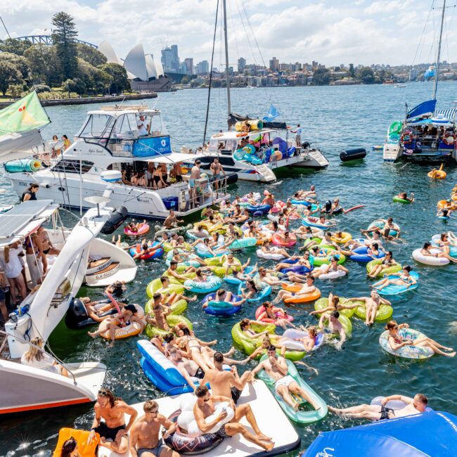 A lively scene of people on inflatable pool floats gathering between several boats on a sunny day in a bay. The city skyline and iconic structures are visible in the background. The atmosphere is festive and crowded.