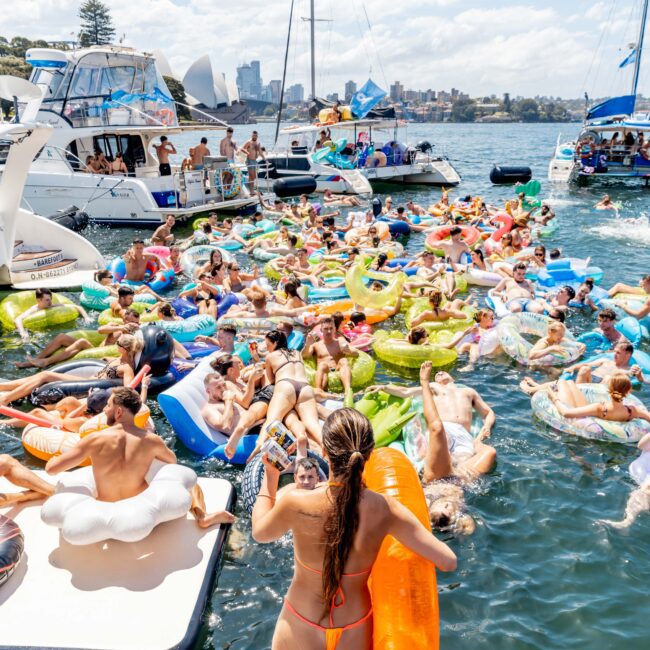 Group of people relaxing on colorful floaties in the water, surrounded by boats. A sunny day with a view of the city skyline in the background. A person in a red swimsuit stands at the edge of a boat holding an orange floatie.