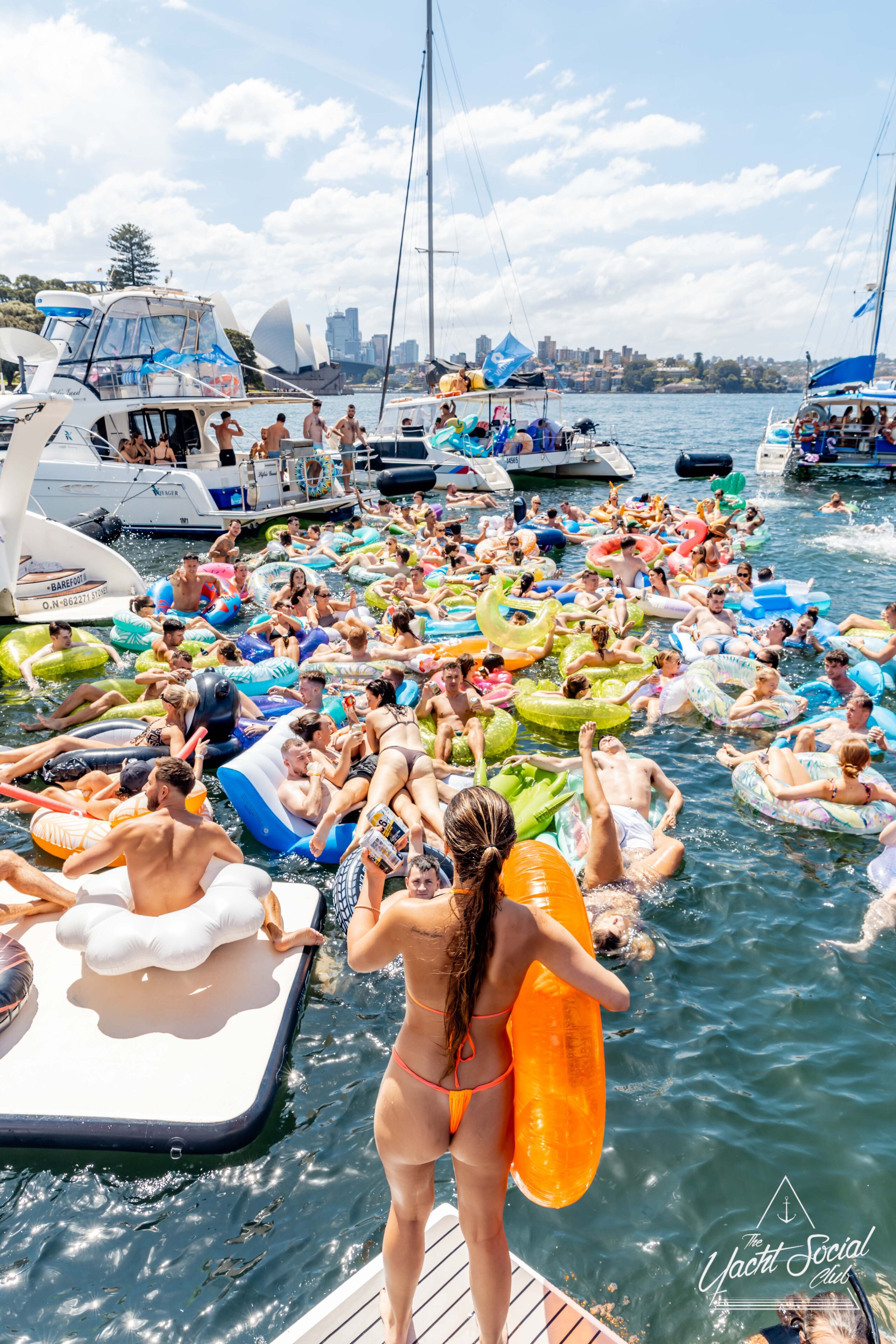 Group of people relaxing on colorful floaties in the water, surrounded by boats. A sunny day with a view of the city skyline in the background. A person in a red swimsuit stands at the edge of a boat holding an orange floatie.