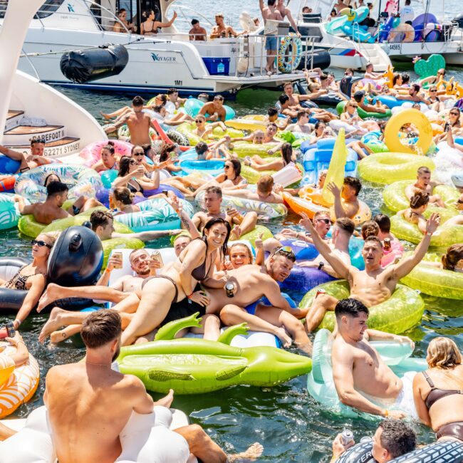 A lively pool party scene with numerous people enjoying the water on colorful floaties. There's a mix of adults and children, surrounded by boats in the background and a city skyline visible in the distance.