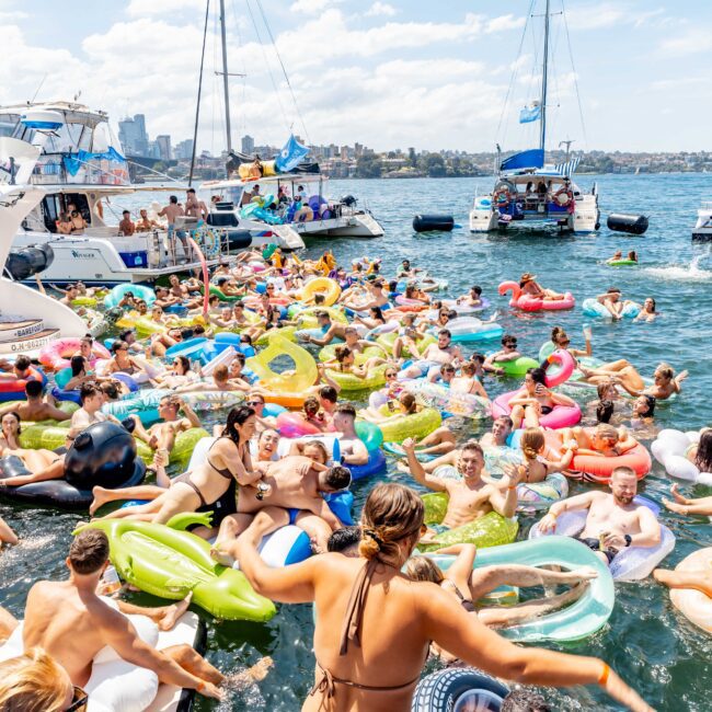 A crowded scene on the water with numerous people on inflatables of various shapes and colors. Boats are anchored nearby, and many participants are enjoying the sunny day. The atmosphere is lively and festive.