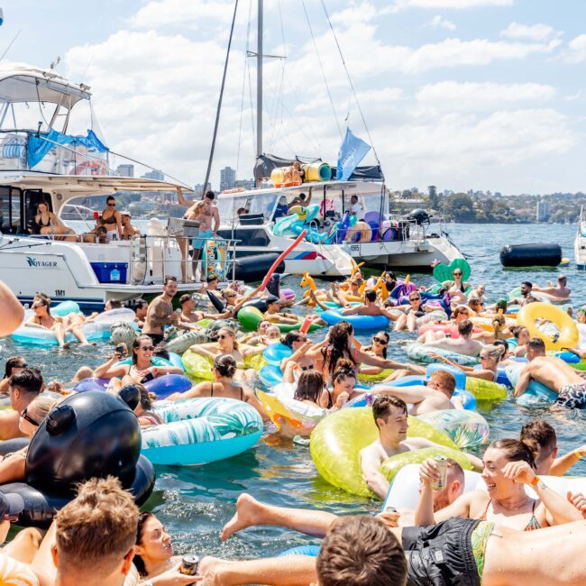 A lively scene of people gathering on colorful inflatables in a sunny, crowded waterway with boats in the background. The atmosphere is festive, with many individuals relaxing and enjoying the day.