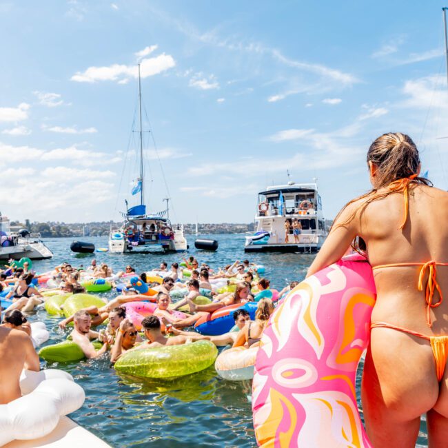 A group of people enjoying a sunny day on a lake, floating on colorful inflatable tubes. A woman in an orange bikini stands in the foreground on a boat, looking towards the water. Sailboats and a motorboat are in the background.