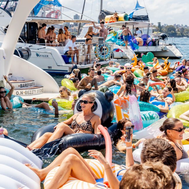 A crowded, lively yacht party on a sunny day. People are enjoying themselves in the water surrounded by inflatable floats of various shapes and colors. Yachts are docked nearby, and partygoers are mingling on the boats and in the water.