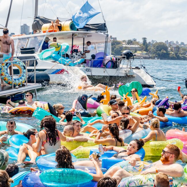 A crowded scene of people relaxing on colorful inflatable pool floats in the water near a docked boat. Many are sunbathing, chatting, or holding drinks, with bright skies and trees in the background. The atmosphere is lively and festive.