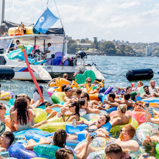 A group of people relax on colorful inflatable floaties in a sunny, crowded harbor. They are near a sailboat, enjoying the water, socializing, and celebrating. The sky is clear, and everyone appears to be having fun.