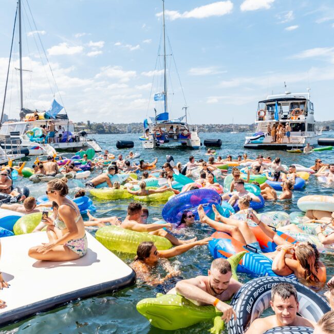 A group of people enjoying a sunny day on the water with inflatable floats of various colors and shapes. Several boats are anchored nearby under a partly cloudy sky. The scene is lively and filled with people relaxing and socializing.