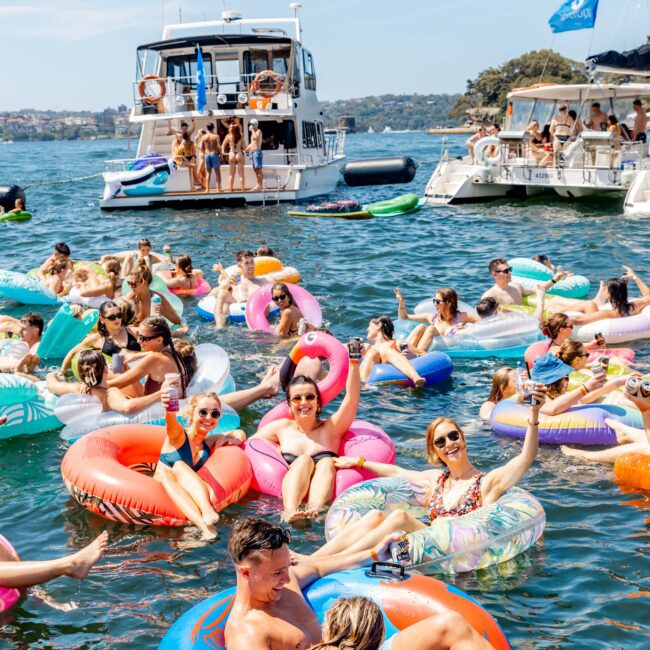 People float on colorful inflatables, enjoying a sunny day on the water. Two boats are in the background, each filled with more people celebrating. Everyone appears relaxed, wearing swimwear and sunglasses, with a festive atmosphere.