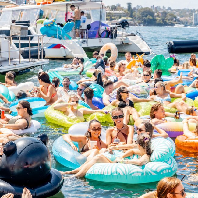 A lively scene of people in swimwear enjoying a sunny day on colorful inflatable floats in the water. A nearby boat is anchored, with a few more floats on its deck. The background shows a distant shoreline and clear skies.