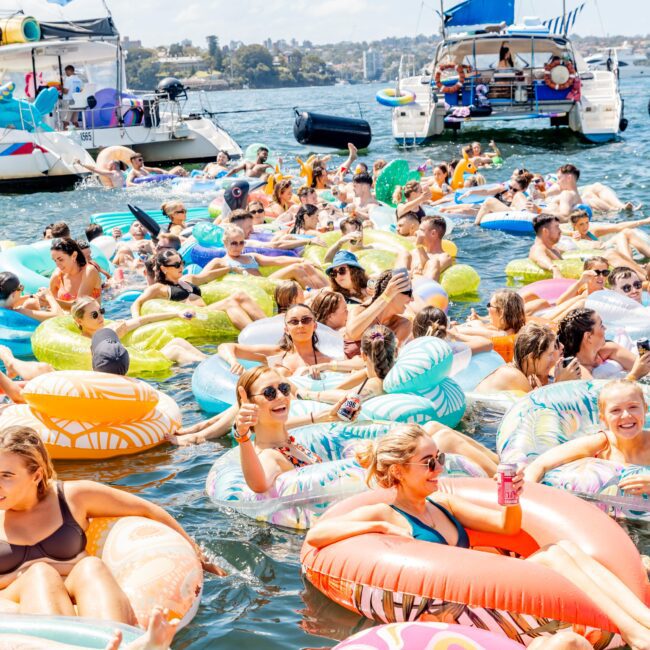 People relax in colorful inflatable rings and floats on a sunny day in a busy body of water. Two boats with sails are anchored nearby. The atmosphere is lively and festive, with everyone enjoying the sun and water.