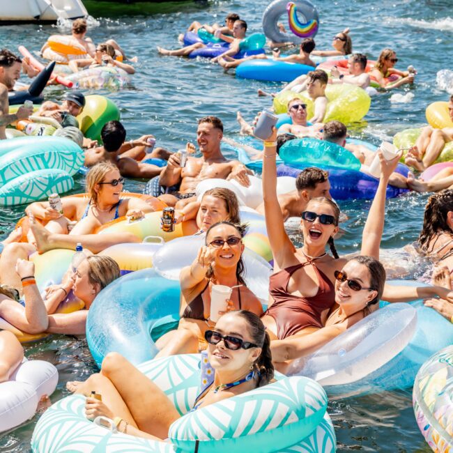 A lively group of people enjoying a sunny day, floating on colorful inflatables in the water. Some are holding drinks and cheering. Boats can be seen in the background with a clear blue sky above.