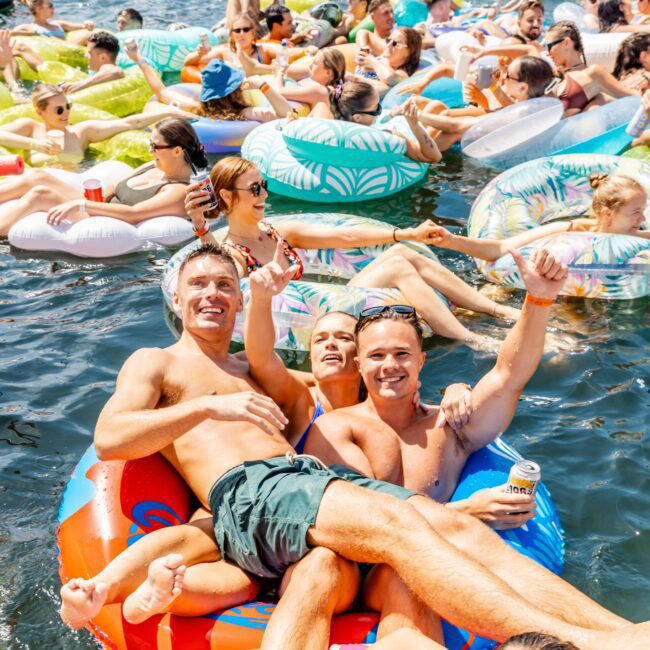 A large group of people enjoying a sunny day on the water, lounging on colorful inflatable floats. They are smiling, relaxing, and holding drinks. Boats are visible in the background, adding to the festive atmosphere.