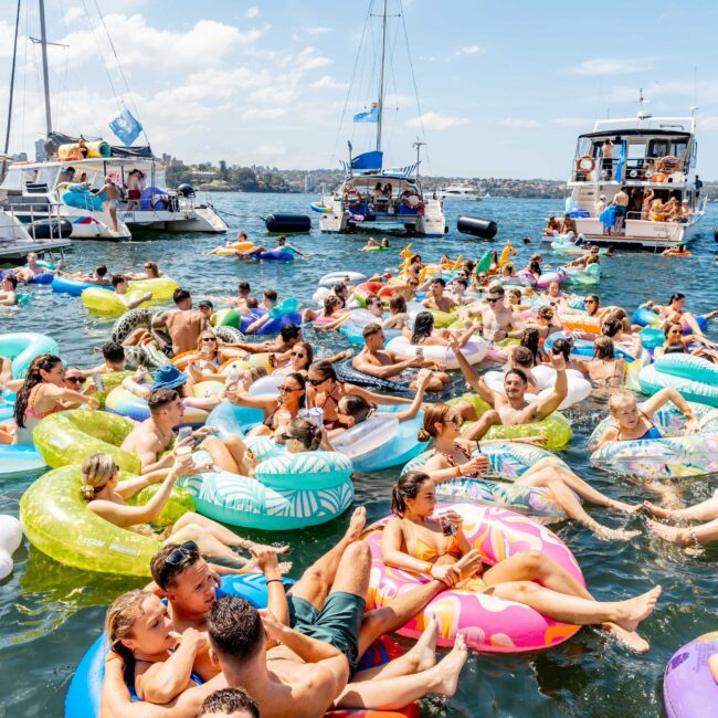 A crowded scene of people relaxing on colorful inflatables in the water, surrounded by several boats. The sky is clear and blue, indicating a bright, sunny day. There is a festive atmosphere as people enjoy the outdoor gathering.