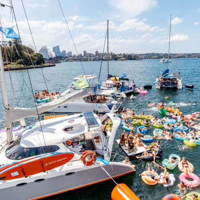 Aerial view of a lively boat party in a harbor, with numerous people on colorful inflatables in the water. Several boats are anchored nearby, and a city skyline is visible in the background.