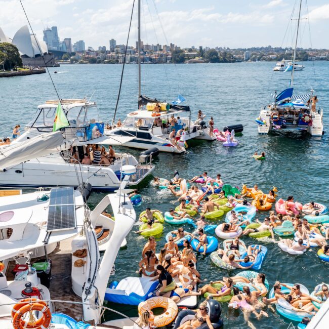 Boats and people on inflatable floats gather in a busy harbor scene. The sky is partly cloudy, and a city skyline is visible in the background. People are enjoying the sunny day on the water, mingling and relaxing.