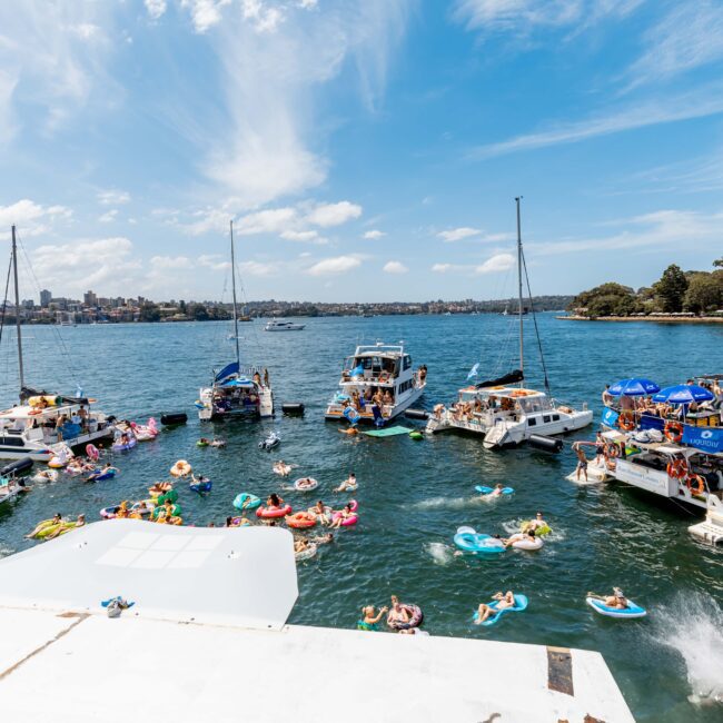 People enjoying a sunny day on a bay with boats anchored close together. Many are swimming and floating on inflatables in the water. The shoreline and city skyline are visible in the background under a blue sky with clouds.