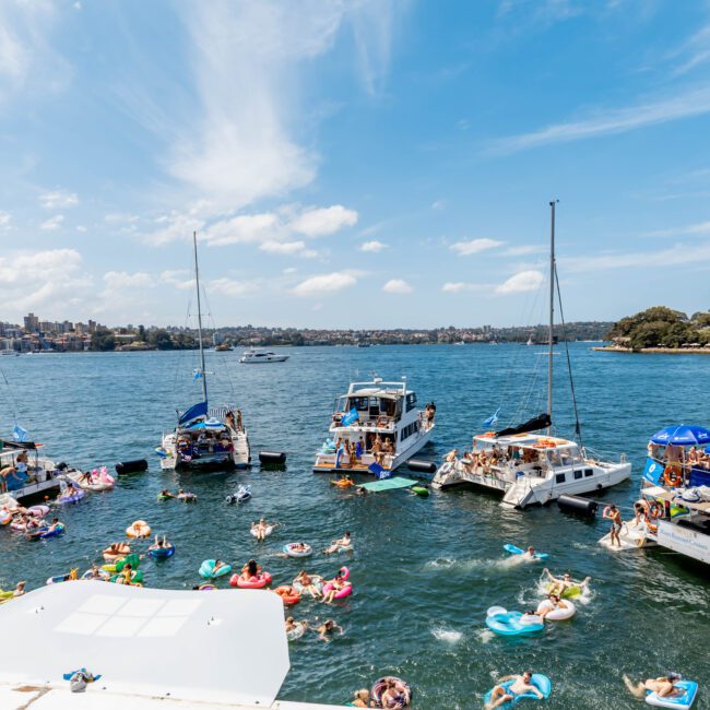 A lively scene on a sunlit harbor with several boats anchored around a group of people enjoying the water on inflatables. The city skyline and an iconic opera house are visible in the background under a partly cloudy sky.