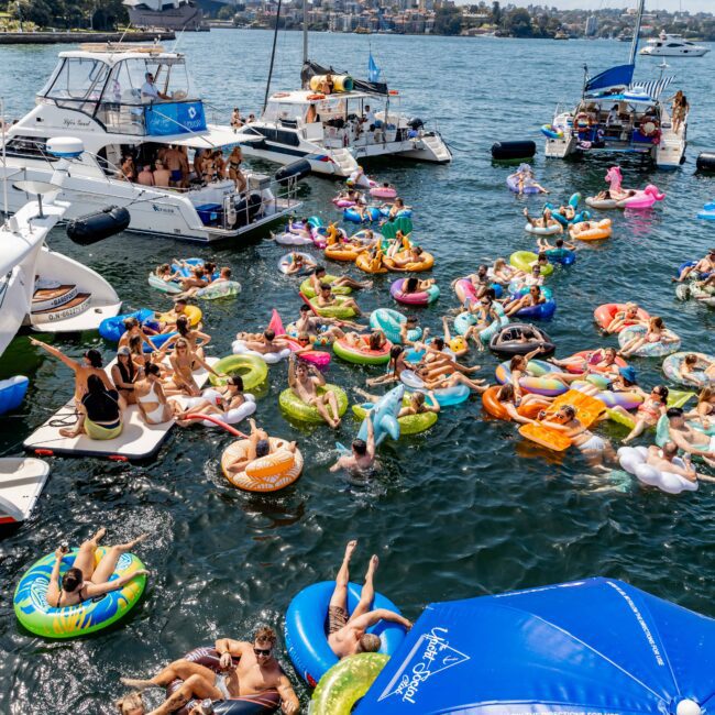 A lively scene of people enjoying a day on a harbor, lounging on colorful inflatables and swimming near several anchored boats. A sunny sky and city skyline are visible in the background, while a large blue umbrella shades part of the dock.