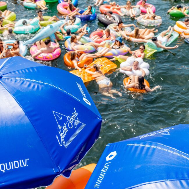 People on colorful inflatable tubes and floats enjoy a sunny day on the water, surrounded by numerous beach umbrellas. The scene is lively, with some floats shaped like animals and visible logos on the umbrellas reading "The Yacht Social Club.