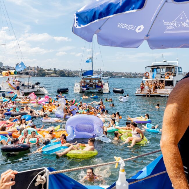 People enjoying a sunny day floating on inflatable toys in the water near docked boats. A large umbrella provides shade in the foreground, and a bottle of sanitizer is visible. The sky is clear, and the atmosphere is festive and relaxed.