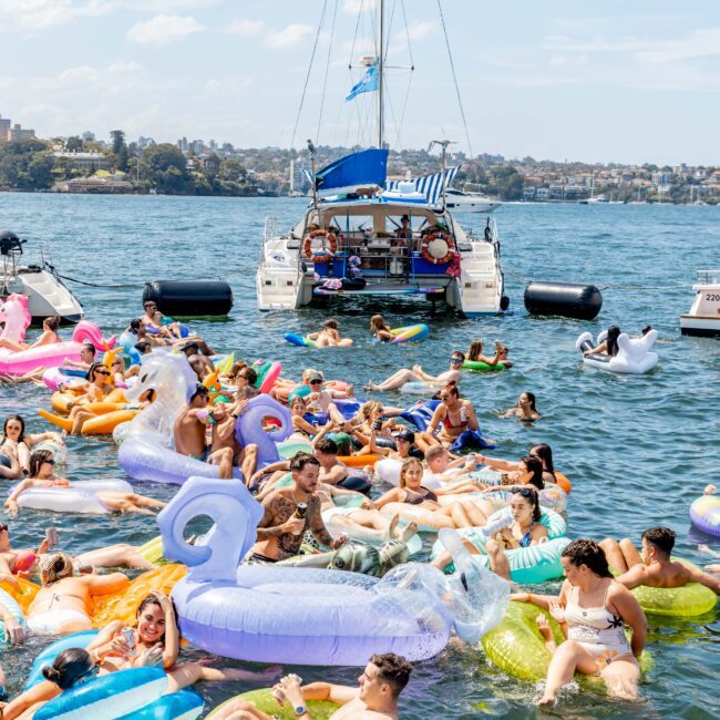 A group of people enjoying a sunny day on colorful inflatable tubes in the water. A boat is anchored nearby, and the shoreline with greenery is visible in the background. The atmosphere is lively and festive.