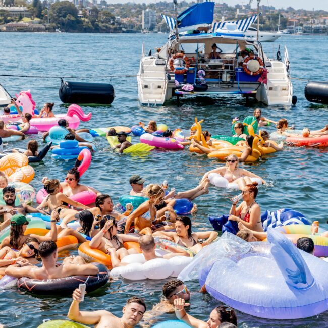 A large group of people enjoy a sunny day on a lake, floating on colorful inflatables of various shapes. A boat is anchored nearby, and the shoreline with a cityscape is visible in the background.