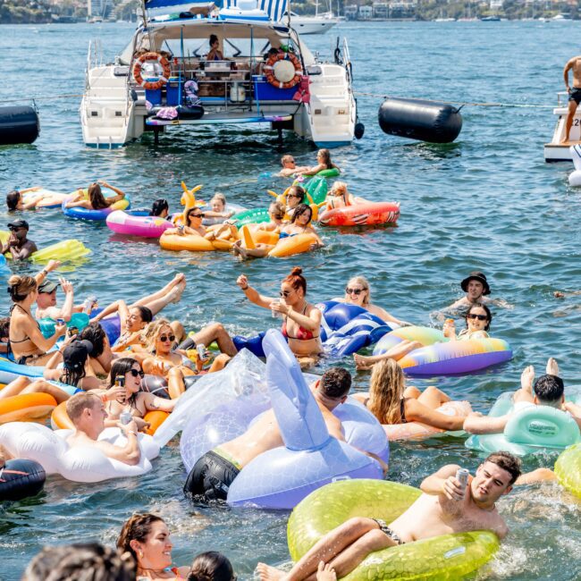 People relaxing on colorful inflatable floaties in the water near a catamaran with flags. The scene is festive with clear skies, and everyone appears to be enjoying a sunny day on the water.