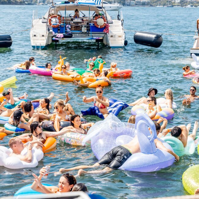 People enjoying a sunny day on a lake, floating on colorful inflatables. A catamaran with a blue canopy is docked nearby. The scene is lively with many people socializing and relaxing in the water.