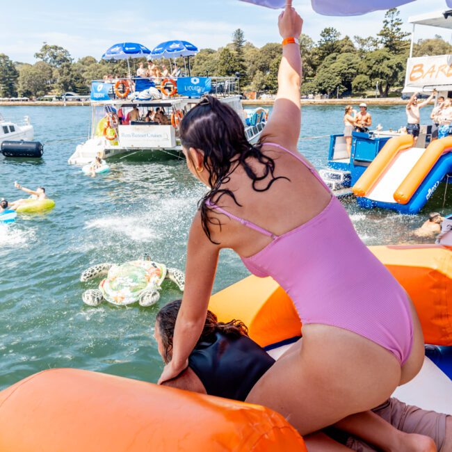 A person in a pink swimsuit assists another into the water from a boat with inflatable slides. People are enjoying the sunny day, some floating on inflatables. Nearby boats have blue umbrellas, and trees line the distant shore.