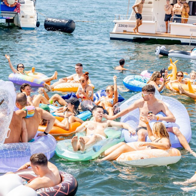 A large group of people enjoying a sunny day on the water, relaxing on colorful inflatable floats near a boat. The scene is lively, with individuals chatting and sunbathing, surrounded by the sparkling blue water and boats in the background.