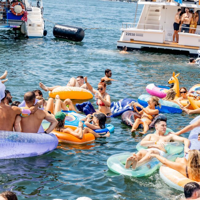 A group of people enjoying a sunny day, floating on colorful inflatable rafts in the water near a yacht. The scene is lively with various inflatables, including a flamingo. Two boats are visible in the background.