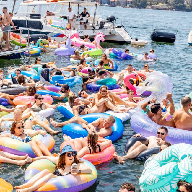 People relax on colorful inflatable pool floats in a crowded water scene by a few anchored boats. The floats include unicorns, flamingos, and geometric shapes. The background shows a calm body of water and a distant city skyline under a partly cloudy sky.