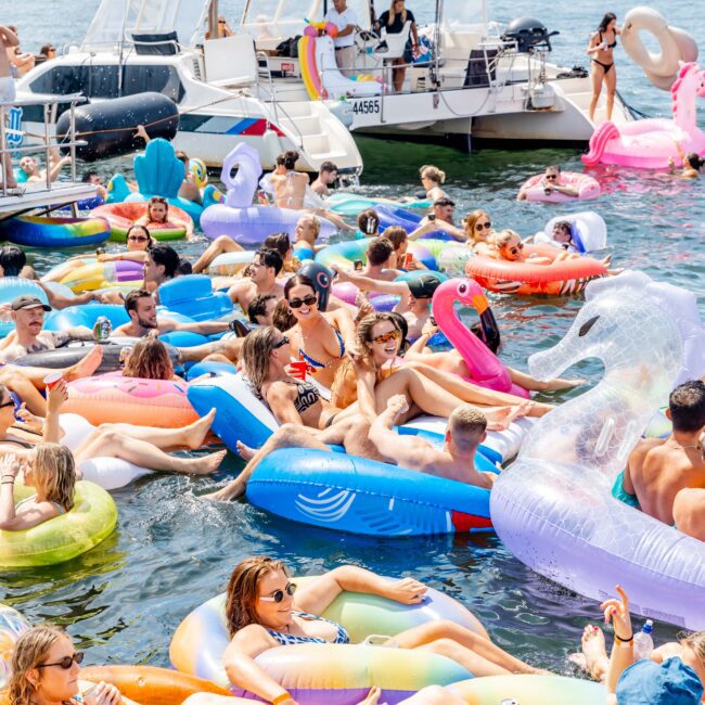 A lively scene on the water with numerous people relaxing on colorful inflatable floats and enjoying themselves. A yacht is visible in the background under a sunny sky.